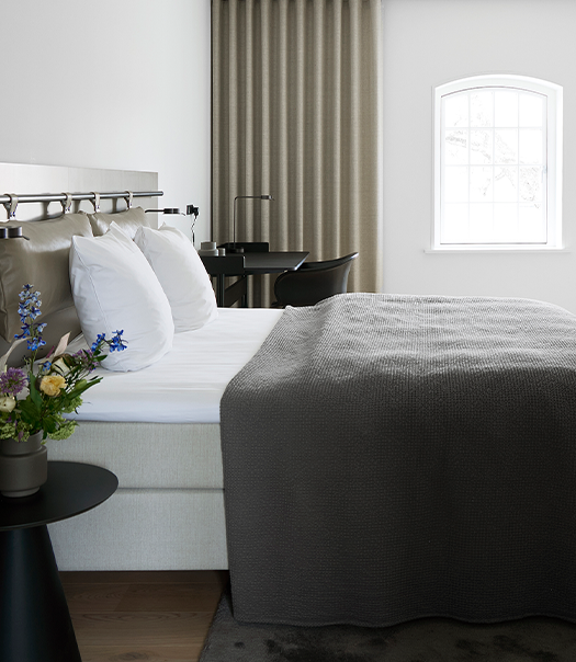 Lyst hotelværelse med dobbeltseng, hvide vægge og blomster på natbord